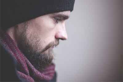 A bearded man with a beanie and scarf sits in profile. He appears sad.